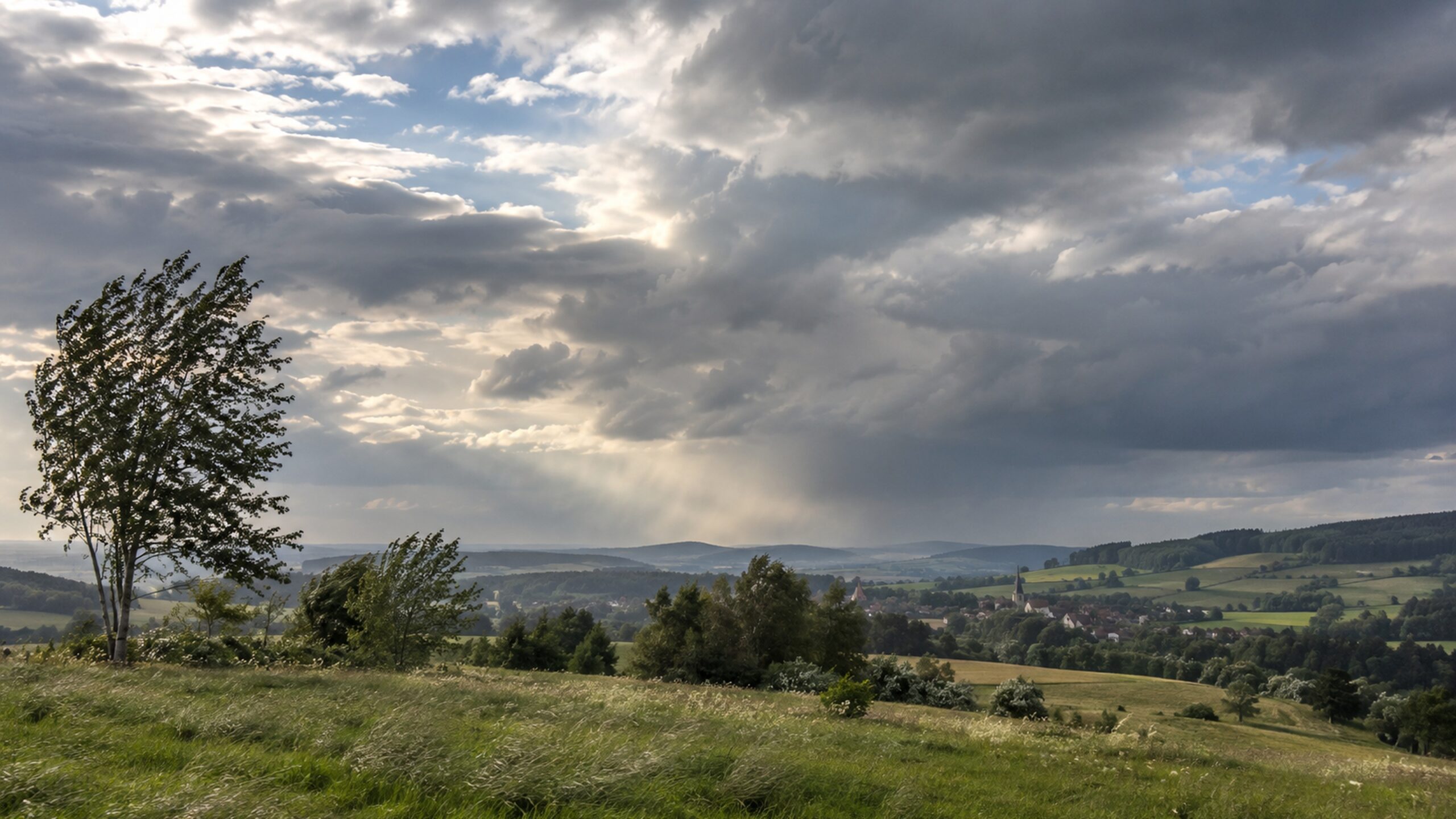 Windige und wechselhafte Wetterlage über einer deutschen Hügellandschaft mit Dorf unter dichten Wolken.