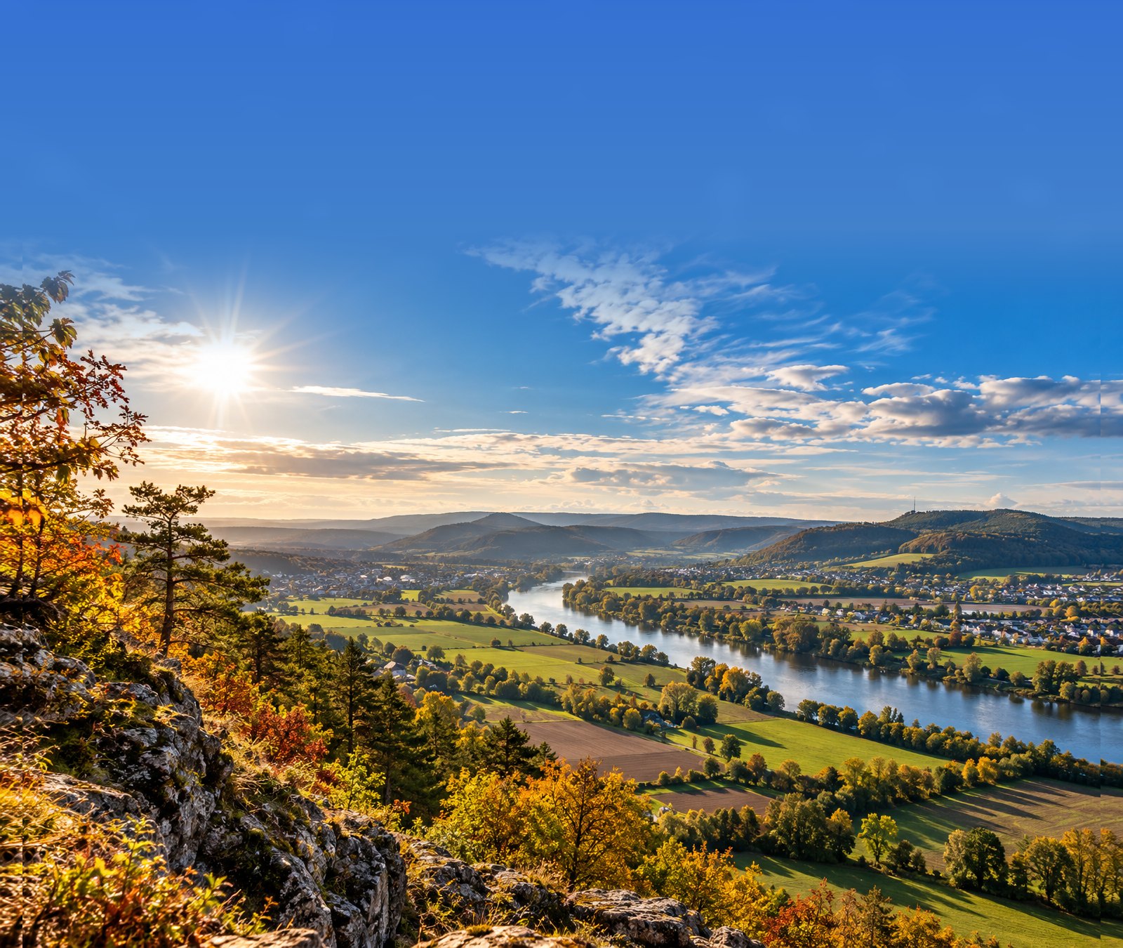 Sonnige spätsommerliche Landschaft in Deutschland mit Feldern, Fluss und herbstlichen Bäumen.