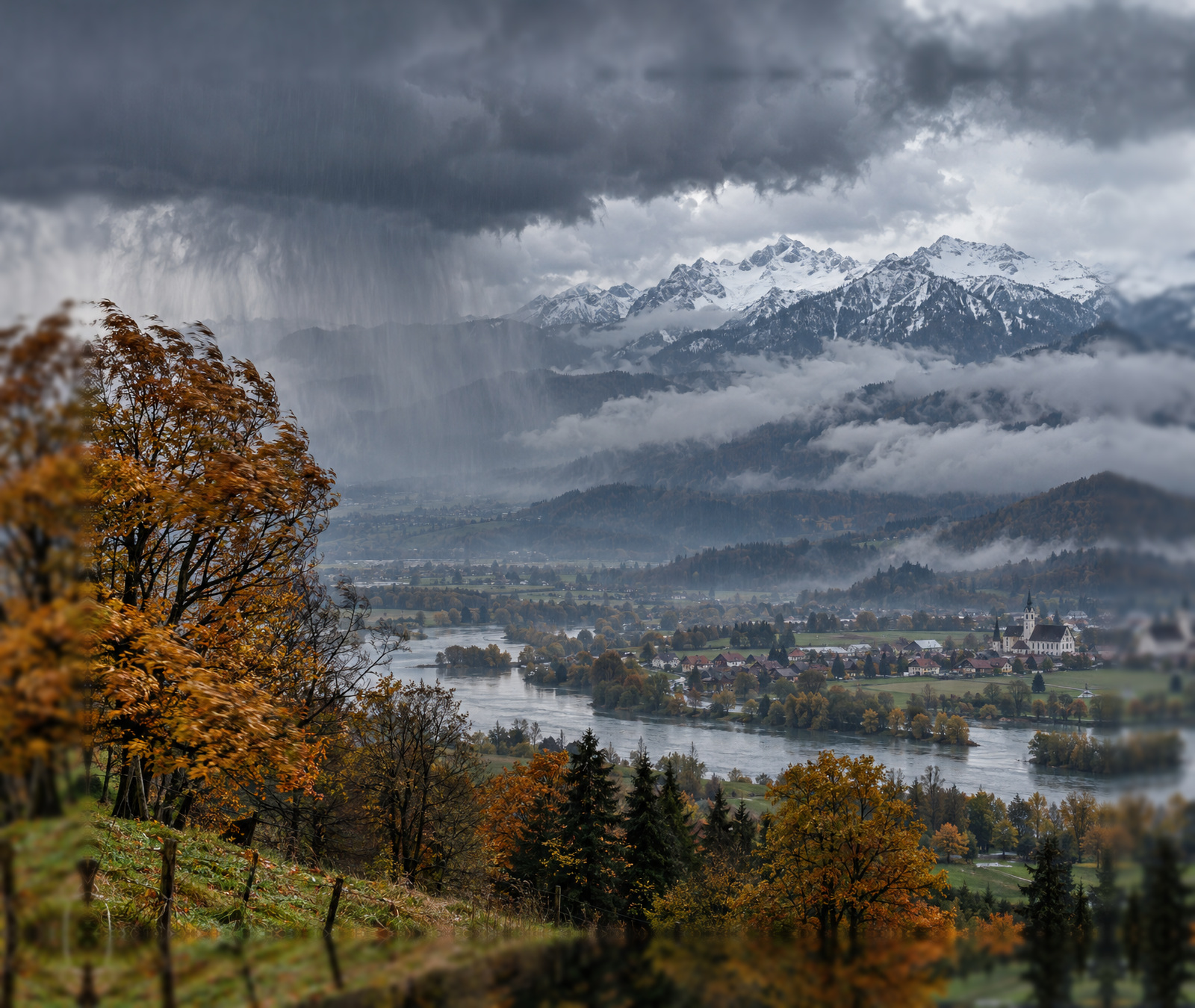 Dunkle Regenwolken über einer herbstlichen Landschaft mit verschneiten Alpen im Hintergrund als Symbolbild für die Kaltfront in Deutschland.