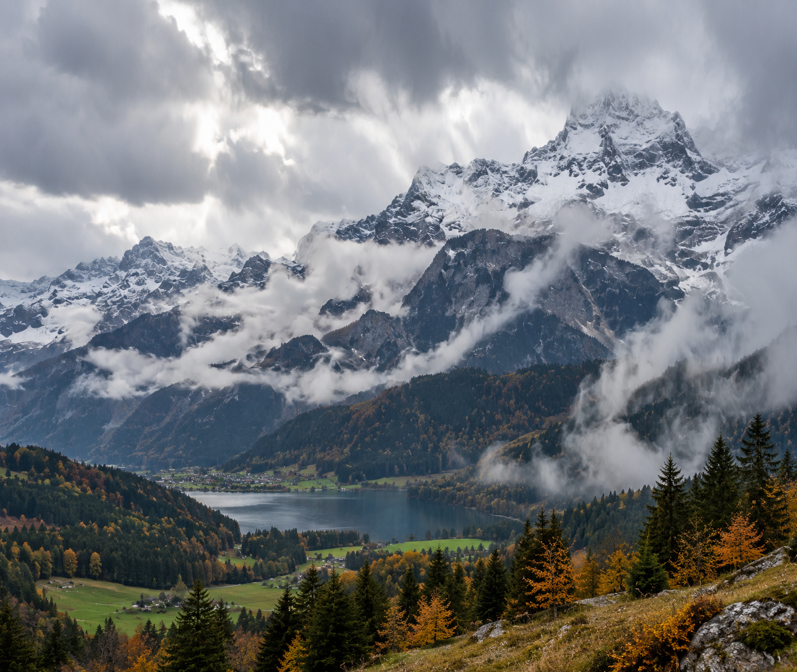 Symbolbild der Bayerischen Alpen mit frisch verschneiten Gipfeln, dunklen Frontwolken und nassen tieferen Hängen.